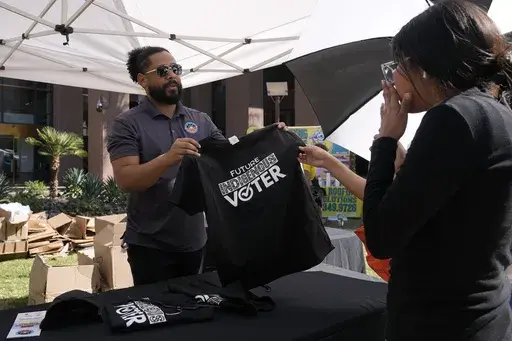 Alexander Castillo-Nunez, left, a civic engagement coordinator at the Inter Tribal Council of Arizona, Inc., shows a voter t-shirt at an Arizona Native Vote booth during an Indigenous Peoples' Day event Monday, Oct. 14, 2024, in Phoenix. (AP Photo/Ross D. Franklin)