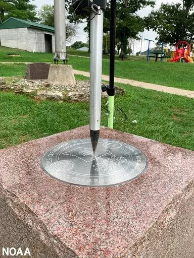 This photo provided by NOAA shows the tip of a tripod resting on the center of the 2020 Center of Population Commemorative Survey mark, as part of a GPS survey to determine the precise latitude, longitude, and height of the mark on Sept. 14, 2022 in Hartville, Mo.  The U.S. Census Bureau announced the nation’s new center of population in 2020 to be in Hartville, in the Ozark Mountains.  (NOAA via AP)