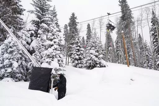 Ian Sidwell adjusts a machine used to make snow at Vail Mountain Resort as snowboarders ride a lift, Wednesday, Dec. 29, 2021, in Vail, Colo. Newer snowmaking technology is allowing ski areas to be more efficient with energy and water usage as climate change continues to threaten snowpack levels. (AP Photo/Brittany Peterson)